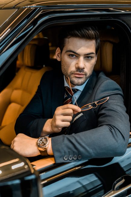 handsome businessman with luxury watch holding eyeglasses while sitting in car